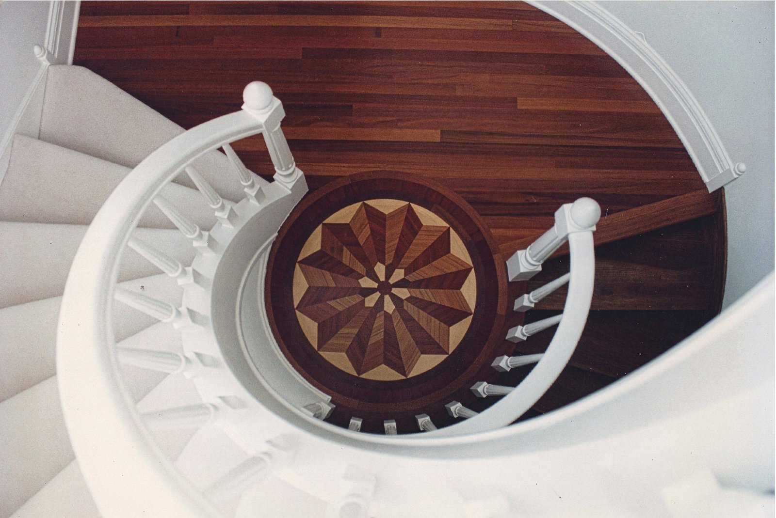 Round starburst medallion set into a dark walnut floor at the foot of a curved staircase — walnut and maple, custom commission